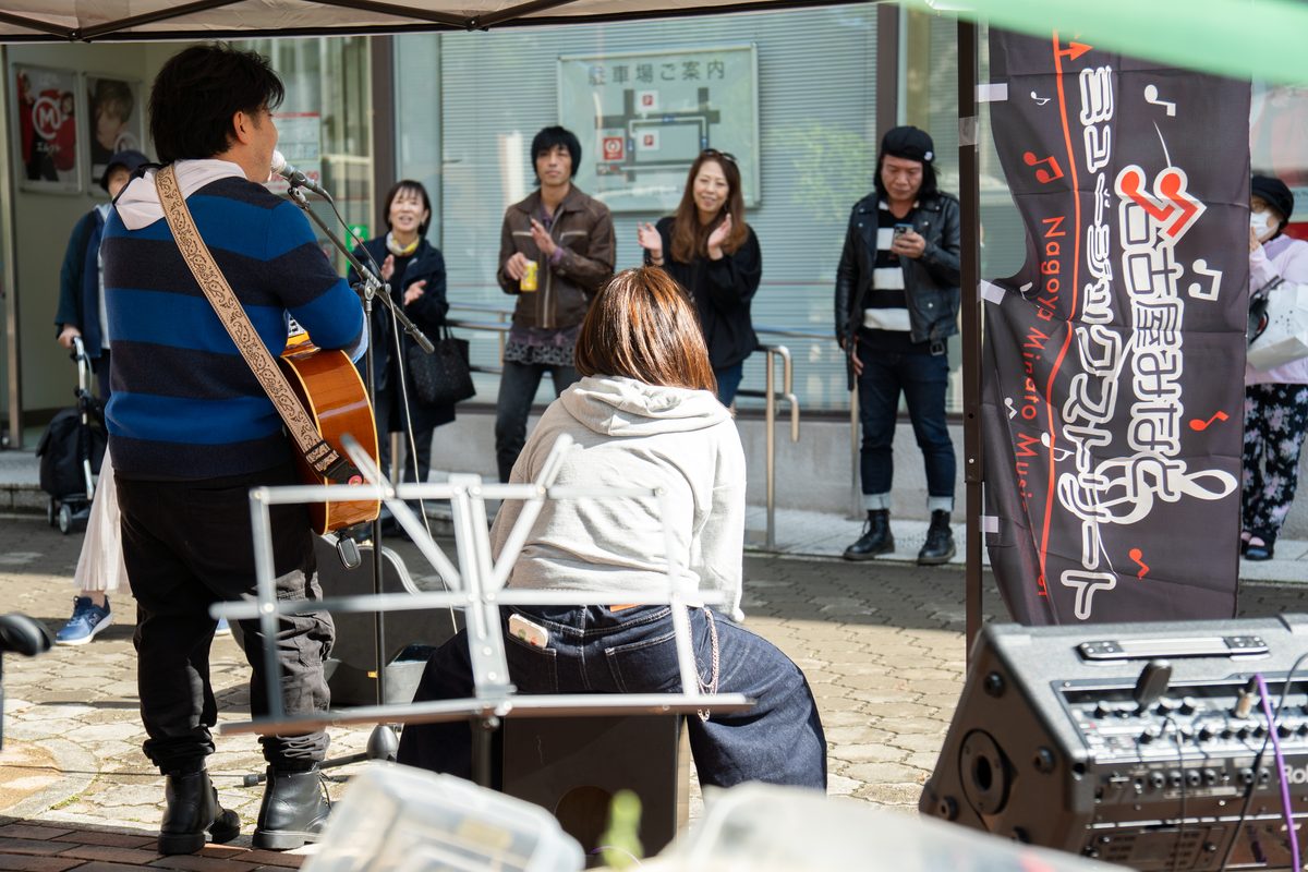 のぼりの前で弾き語る出演者と、拍手を送る観客