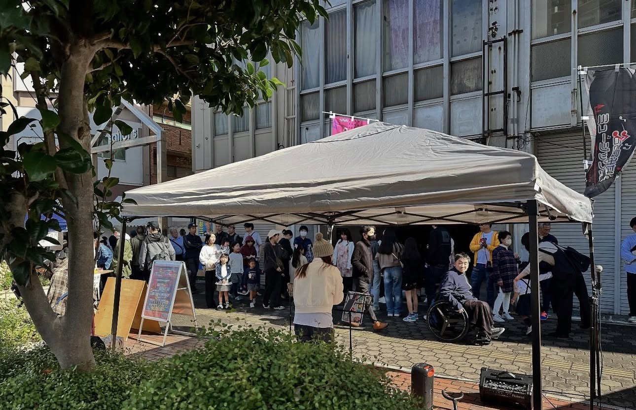 商店街の歩道のテントで演奏する出演者と、集まって聴く家族連れ